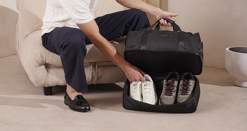 CALPAK Stevyn Duffle in black with bottom zipper shoe compartment that model is placing a pair of white shoes and hiking shoes in